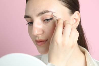 Young woman plucking eyebrow with tweezers on pink background, closeup