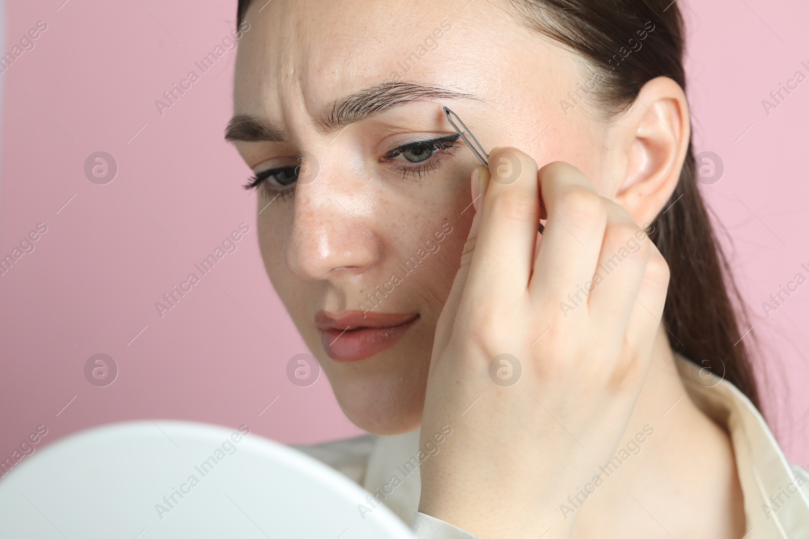 Photo of Young woman plucking eyebrow with tweezers on pink background, closeup