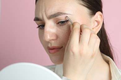 Photo of Young woman plucking eyebrow with tweezers on pink background, closeup