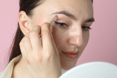 Young woman plucking eyebrow with tweezers on pink background, closeup