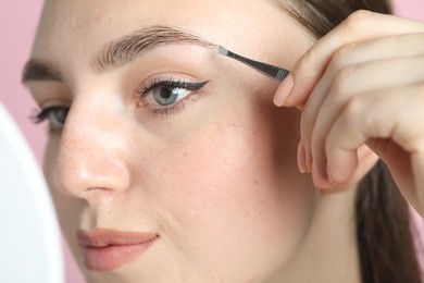 Young woman plucking eyebrow with tweezers on pink background, closeup