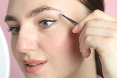 Photo of Young woman plucking eyebrow with tweezers on pink background, closeup