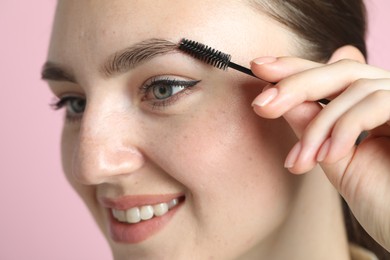 Young woman brushing eyebrow on pink background, closeup