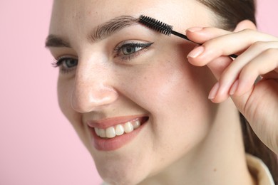 Young woman brushing eyebrow on pink background, closeup