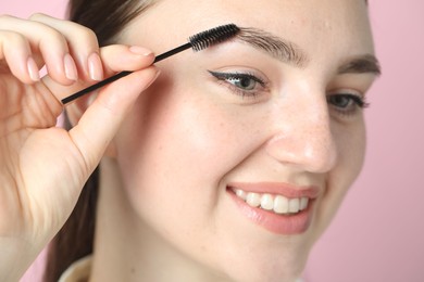 Young woman brushing eyebrow on pink background, closeup