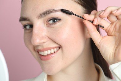 Young woman brushing eyebrow on pink background, closeup