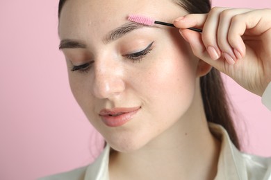 Young woman brushing eyebrow on pink background, closeup