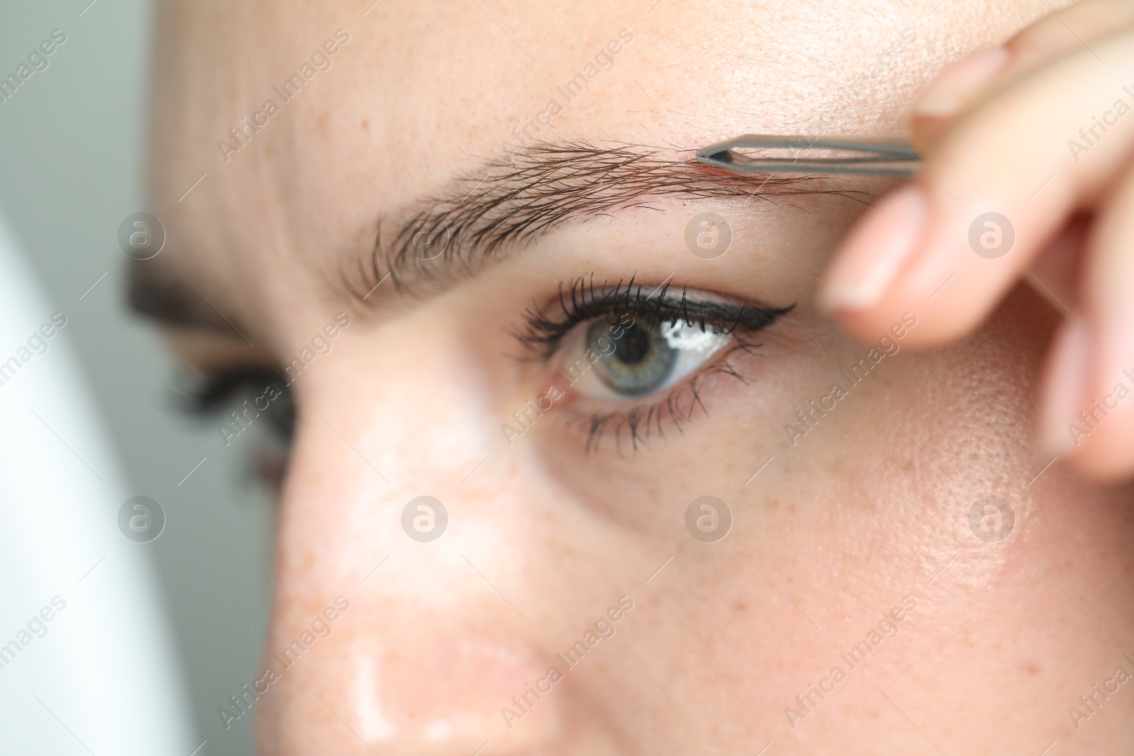 Photo of Young woman plucking eyebrow with tweezers on light background, closeup