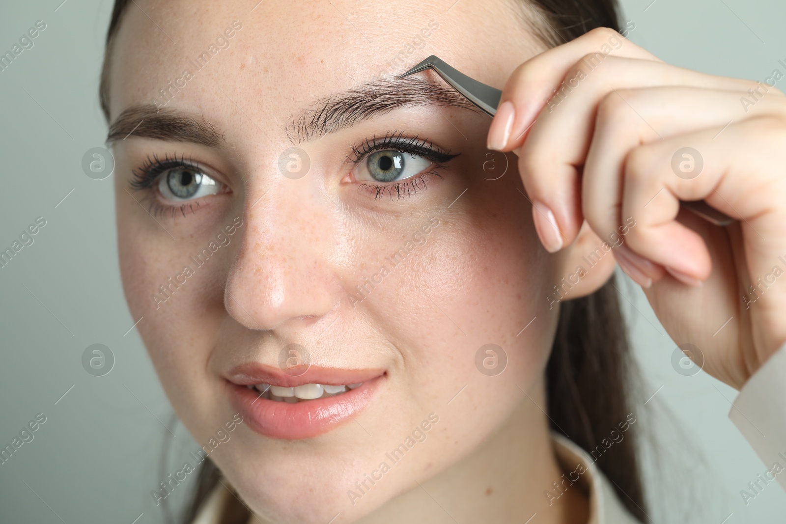 Photo of Young woman plucking eyebrow with tweezers on light background, closeup