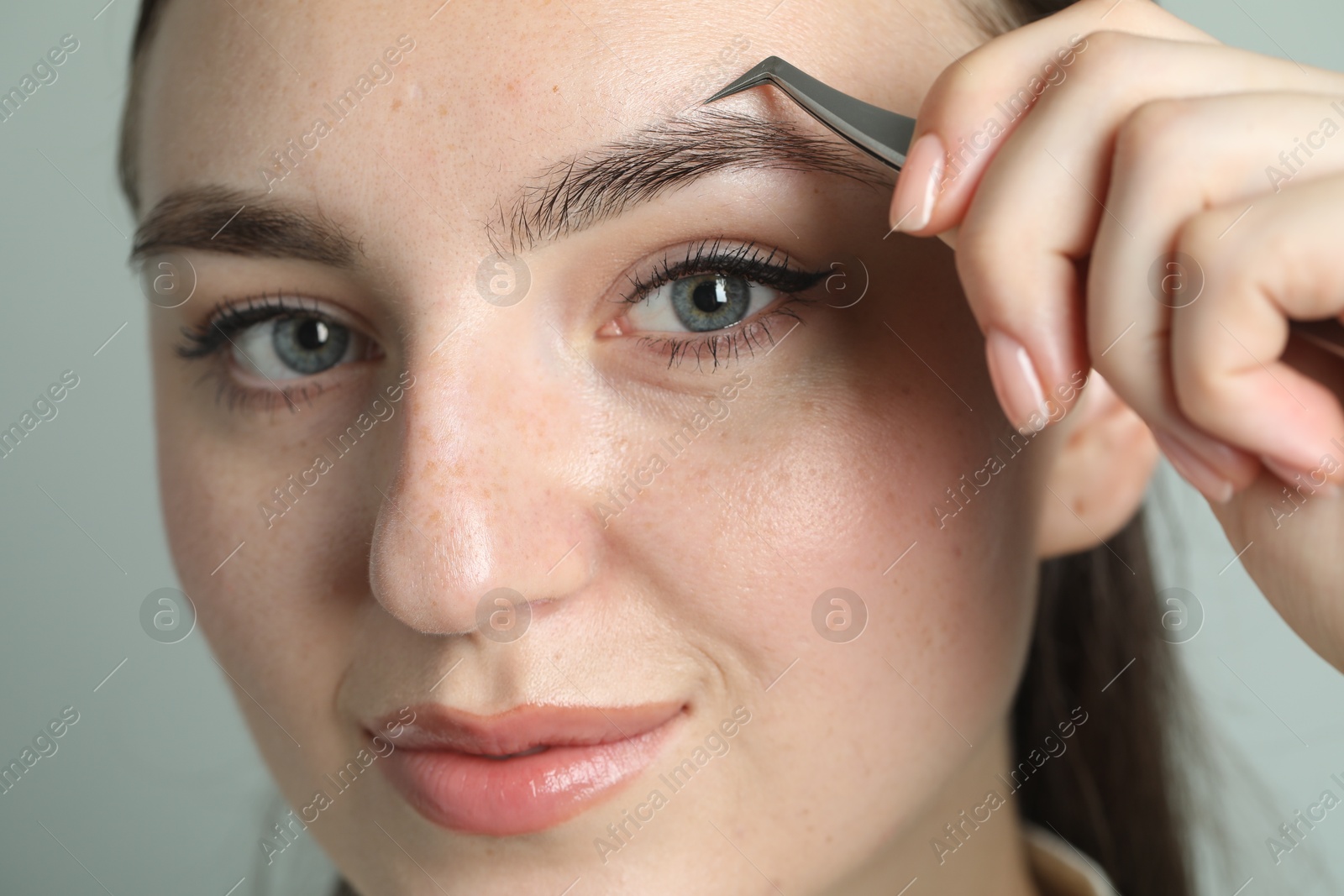 Photo of Young woman plucking eyebrow with tweezers on light background, closeup