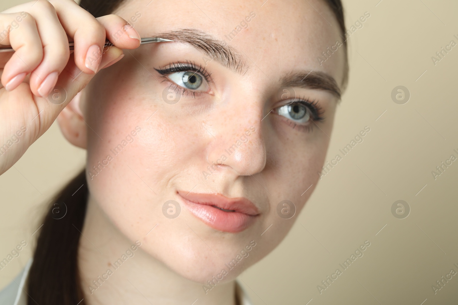 Photo of Young woman plucking eyebrow with tweezers on beige background, closeup