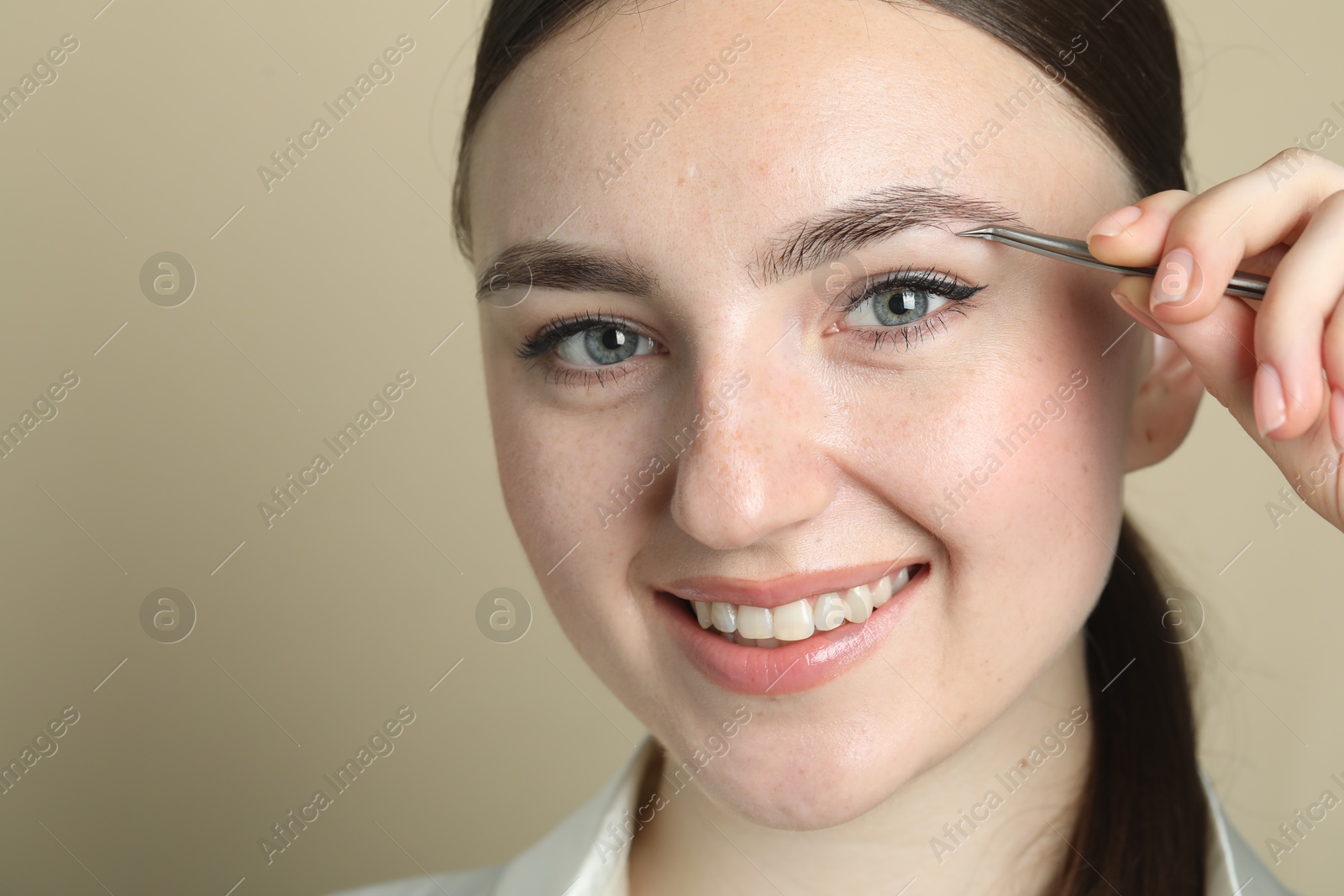 Photo of Young woman plucking eyebrow with tweezers on beige background, closeup. Space for text