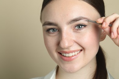 Young woman plucking eyebrow with tweezers on beige background, closeup. Space for text