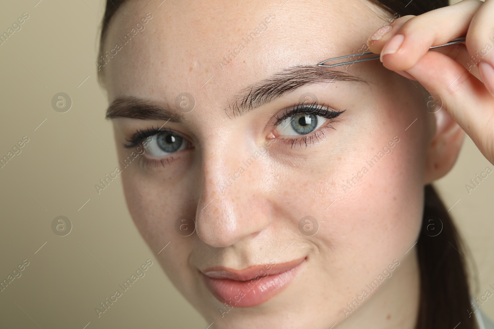 Photo of Young woman plucking eyebrow with tweezers on beige background, closeup