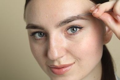 Young woman plucking eyebrow with tweezers on beige background, closeup