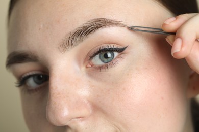 Photo of Young woman plucking eyebrow with tweezers on beige background, closeup