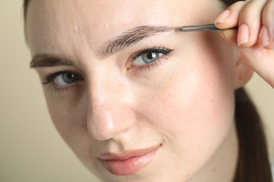 Photo of Young woman plucking eyebrow with tweezers on beige background, closeup