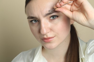 Young woman plucking eyebrow with tweezers on beige background, closeup