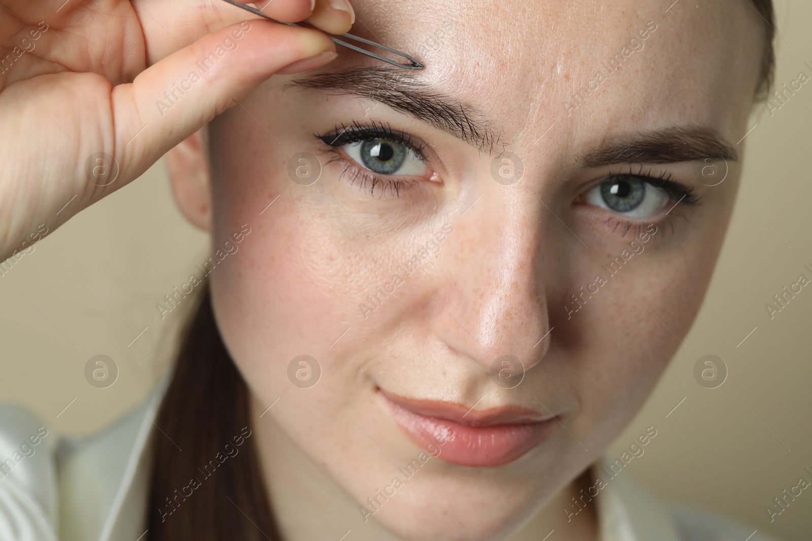 Photo of Young woman plucking eyebrow with tweezers on beige background, closeup