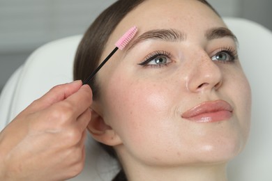 Photo of Beautician brushing young woman's eyebrow in beauty salon, closeup