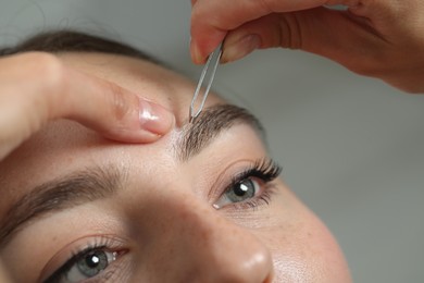 Beautician plucking young woman's eyebrow on light background, closeup