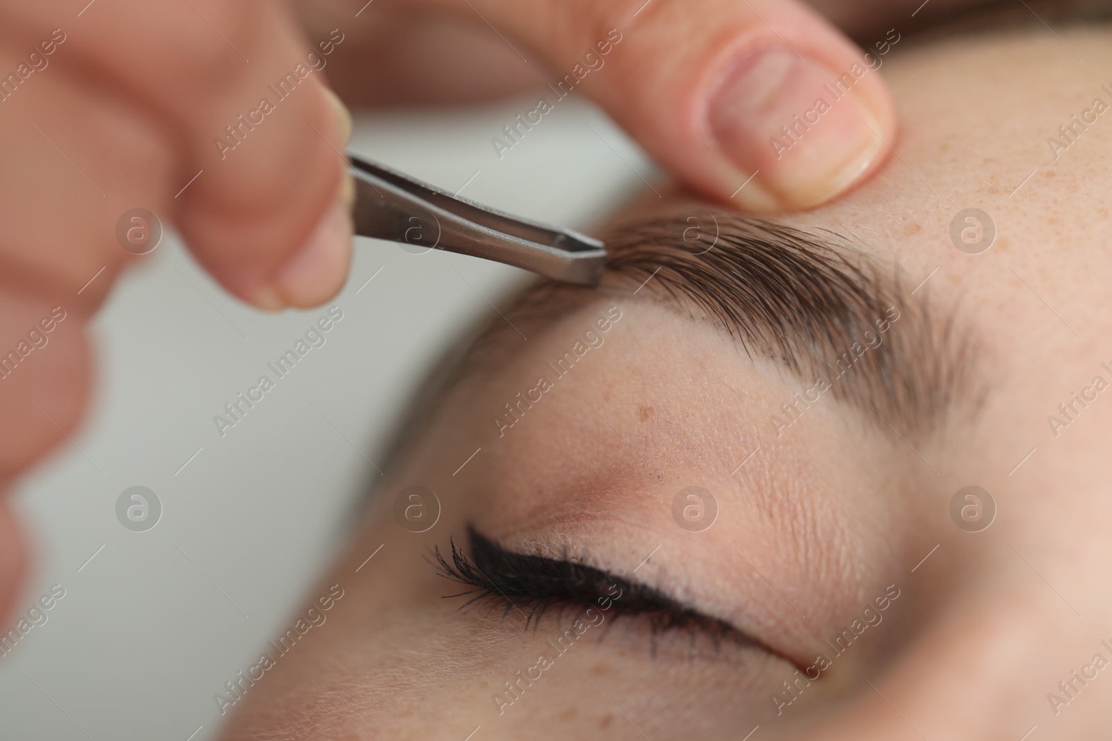 Photo of Beautician plucking young woman's eyebrow on light background, closeup