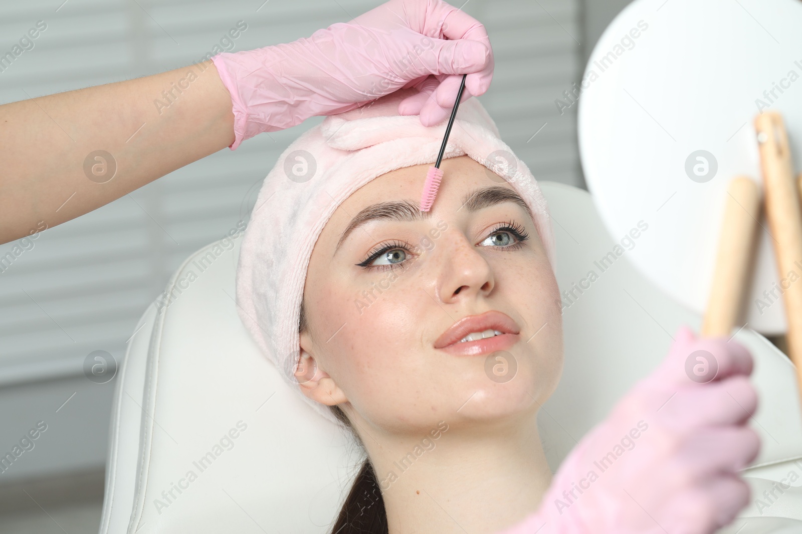 Photo of Beautician brushing young woman's eyebrow in beauty salon, closeup