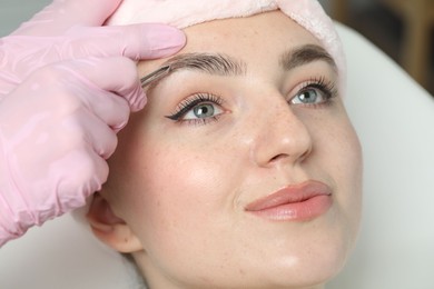 Photo of Beautician plucking young woman's eyebrow in beauty salon, closeup