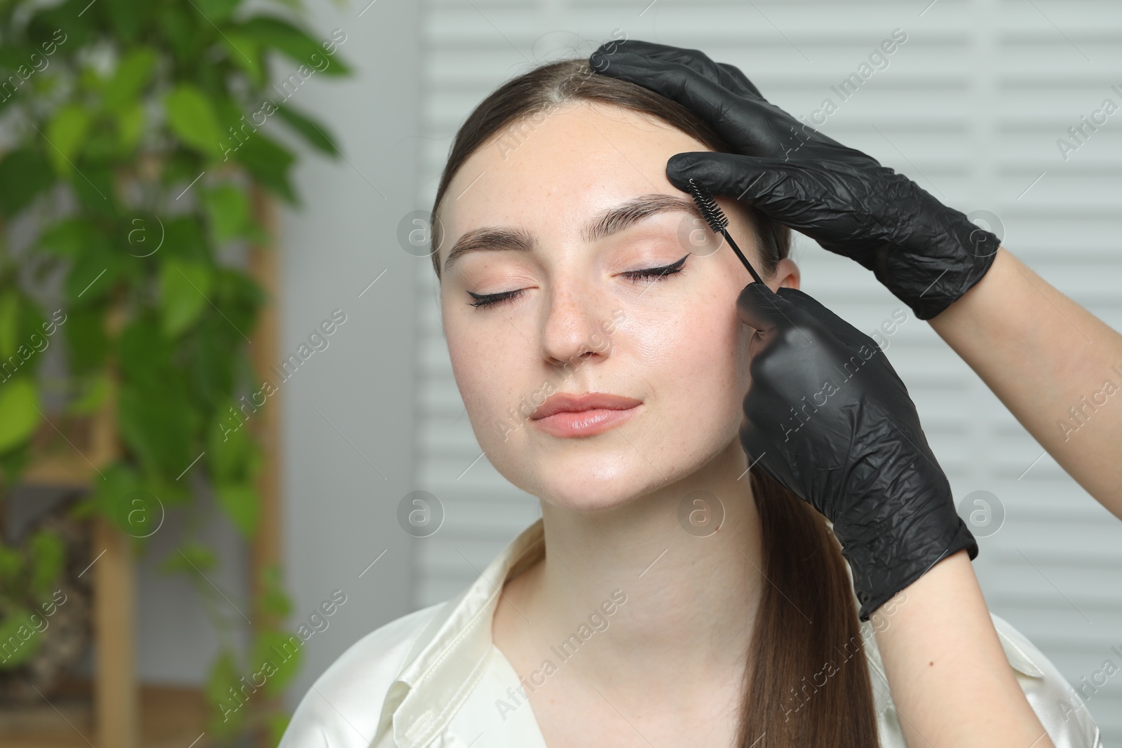 Photo of Beautician brushing young woman's eyebrow in beauty salon, closeup