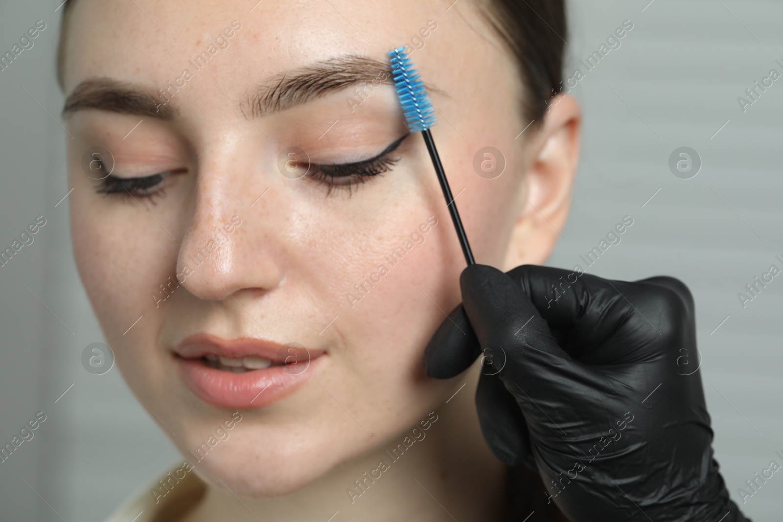 Photo of Beautician brushing young woman's eyebrow in beauty salon, closeup