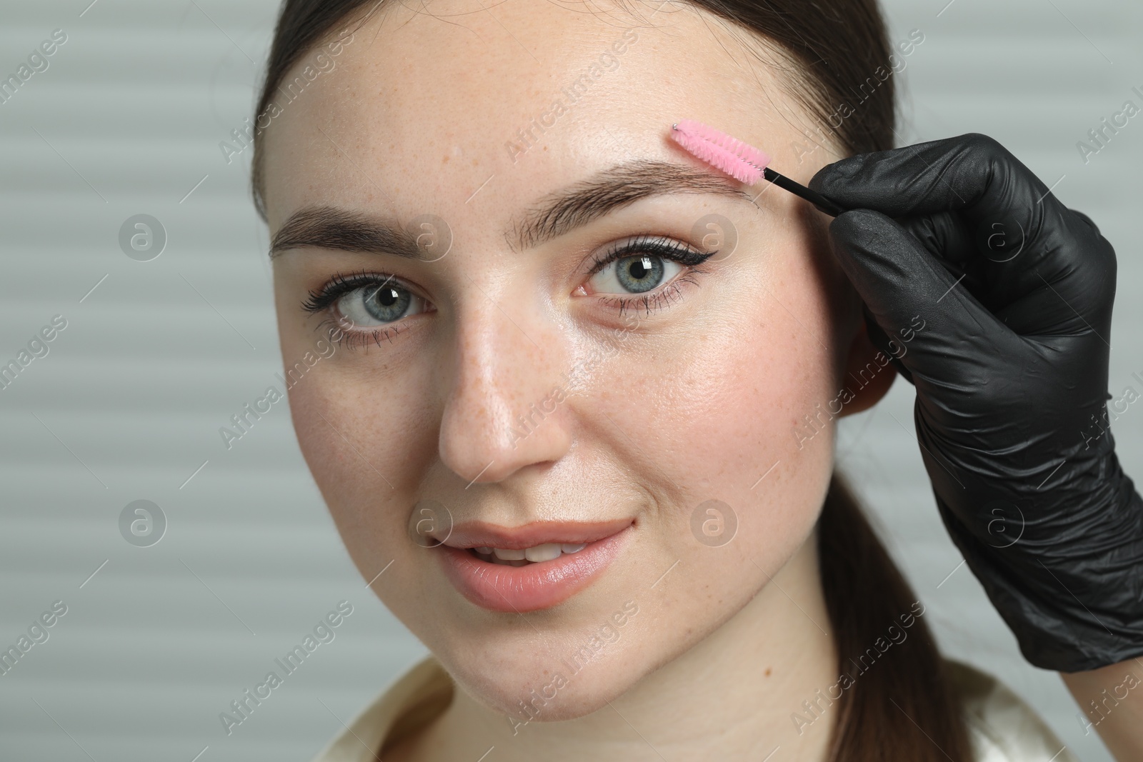 Photo of Beautician brushing young woman's eyebrow in beauty salon, closeup