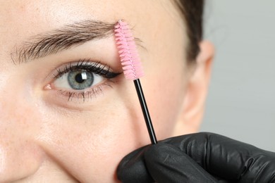 Beautician brushing young woman's eyebrow on light background, closeup