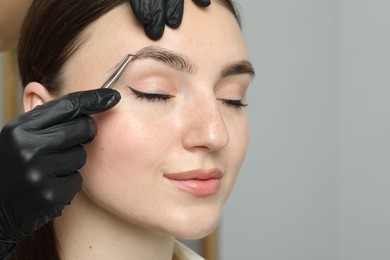 Photo of Beautician plucking young woman's eyebrow on light background, closeup and space for text