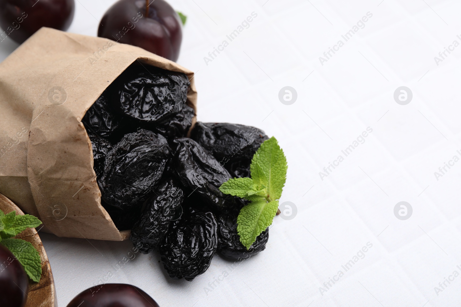 Photo of Paper bag with delicious prunes, fresh ripe plums and mint on white tiled table, closeup. Space for text
