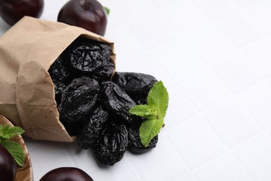 Photo of Paper bag with delicious prunes, fresh ripe plums and mint on white tiled table, closeup. Space for text