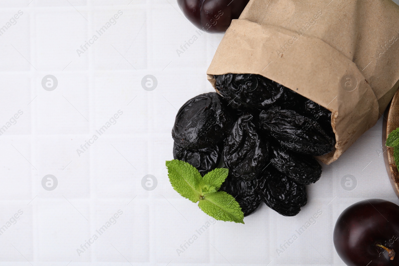 Photo of Paper bag with delicious prunes, fresh ripe plums and mint on white tiled table, flat lay. Space for text