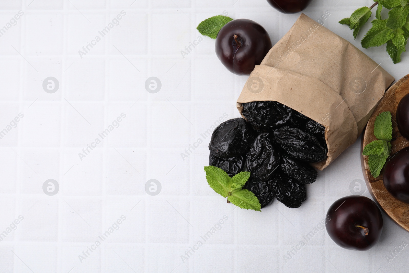 Photo of Paper bag with delicious prunes, fresh ripe plums and mint on white tiled table, flat lay. Space for text