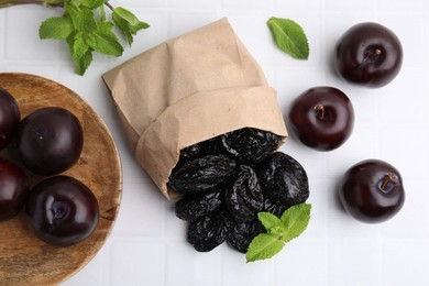 Paper bag with delicious prunes, fresh ripe plums and mint on white tiled table, flat lay