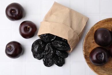 Paper bag with delicious prunes and fresh ripe plums on white tiled table, flat lay