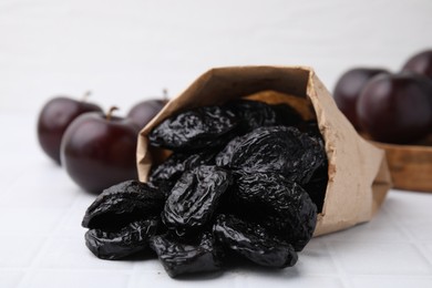 Paper bag with delicious prunes and fresh ripe plums on white tiled table, closeup