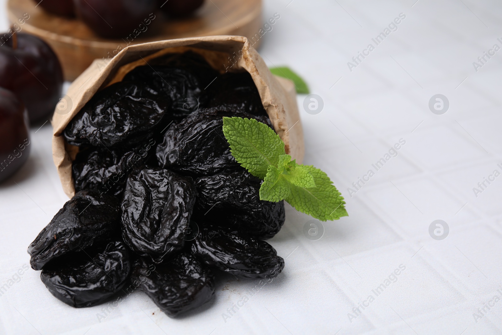 Photo of Paper bag with tasty dried plums (prunes) and mint on white tiled table, closeup. Space for text