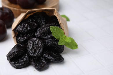 Photo of Paper bag with tasty dried plums (prunes) and mint on white tiled table, closeup. Space for text