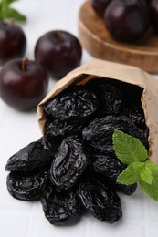 Photo of Paper bag with tasty dried plums (prunes) and mint on white tiled table, closeup