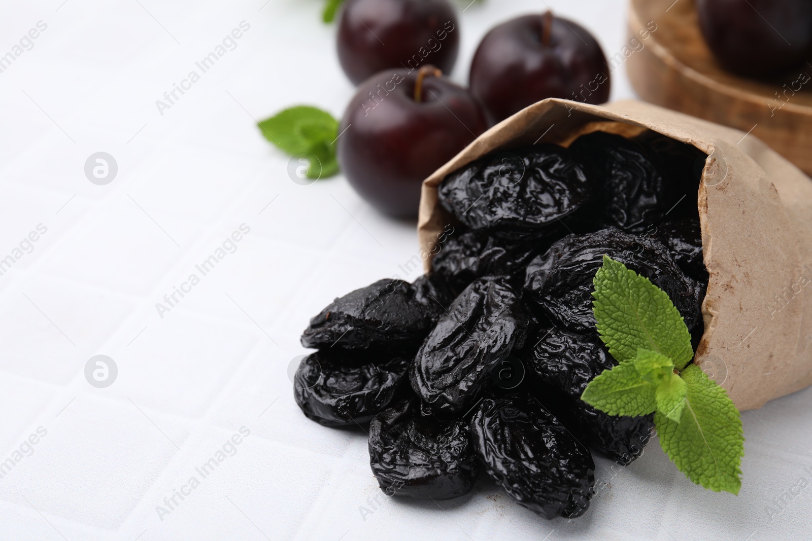 Photo of Paper bag with delicious prunes, mint in and fresh ripe plums on white tiled table, closeup. Space for text