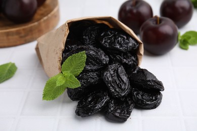 Photo of Paper bag with delicious prunes, mint in and fresh ripe plums on white tiled table, closeup