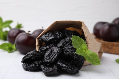 Paper bag with delicious prunes, mint in and fresh ripe plums on white tiled table, closeup