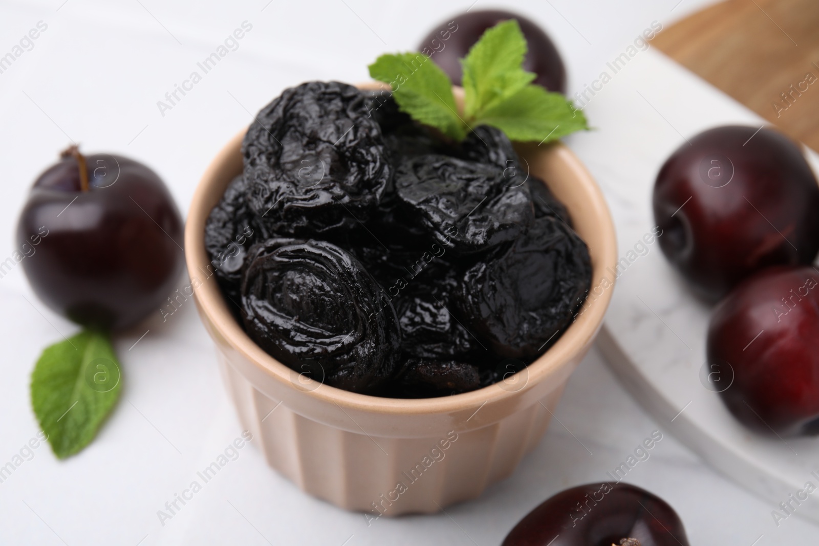 Photo of Delicious prunes with mint in bowl and fresh ripe plums on white table, closeup