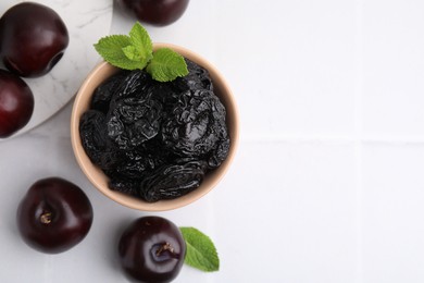 Photo of Delicious prunes with mint in bowl and fresh ripe plums on white tiled table, flat lay. Space for text