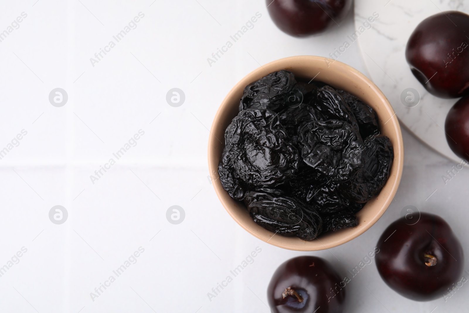 Photo of Delicious prunes in bowl and fresh ripe plums on white tiled table, flat lay. Space for text