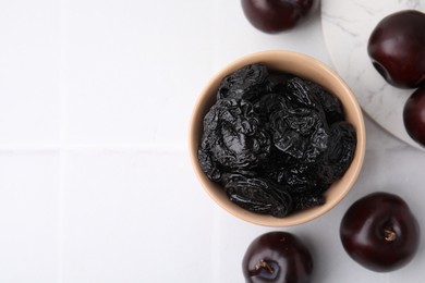 Delicious prunes in bowl and fresh ripe plums on white tiled table, flat lay. Space for text